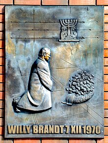 Bronzová plastika zachycující muže klečícího před věncem. Ve spodní části je nápis „Willy Brandt 7 XII 1970“