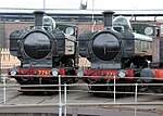 Two pannier tank locomotives are seen from the front and to the right and are standing next to each other in front of a turntable. Both locomotives have black chimneys and fronts. The cabs, pannier tanks, clackbox covers and boiler domes are all green. The buffer beams are red and include the locomotive number in yellow with black edging, 7760 on the left locomotive and 7752 on the right. On both locomotives, one round cab window is visible and is edged in brass.