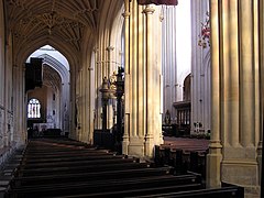 An aisle in Bath Abbey, Bath, England. The aisle is lined with wooden seating (pews), the nave seating can be seen on the right, beyond the arcade pillars. The roof of the aisle is fan-vaulted.