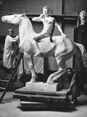 女性彫刻家が馬の彫像の首へ自身の左手を置いている。馬の首は彼女の頭上まで伸びている。