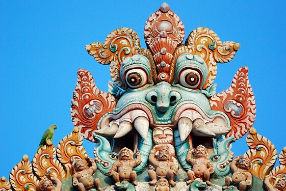 Kleiner Alexandersittich sitzt auf der Schulter des Kirtimukha (Kala) auf der Spitze des Gopuram vom Jambukeshwara-Tempel in Sriramgam, Tamil Nadu, Indien