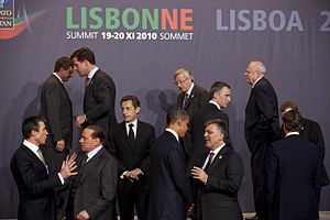Twelve men in black suits stand talking in small groups under a backdrop with the words Lisbonne and Lisboa.