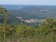 barevná fotografie hlubokého zalesněného údolí pořízená z vysokého bodu.  Skalní výchoz nese hrad a pod lesem prorazí bílý skalnatý bar.