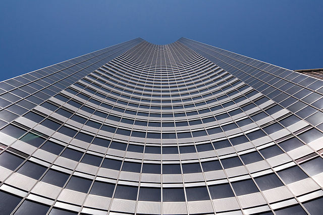 Photographie du bâtiment depuis le bas jusqu'au ciel. Il forme une sorte de gouttière ouverte concave.