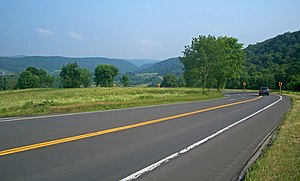 A paved two-lane road crosses the lower half of the picture from left to right. A small blue car is visible at the right with its brake lamps on, where the road turns back sharply. After the turn, it is slightly downhill from the camera and cannot be seen, although black-on-yellow arrow signs indicate where it is. In the distance, in the top of the image, is a valley with green hills on either side