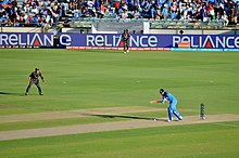 Photographie d'un match de cricket. Sous les yeux de défenseurs, un joueur cherche à frapper la balle alors qu’elle est en train de rebondir au sol.