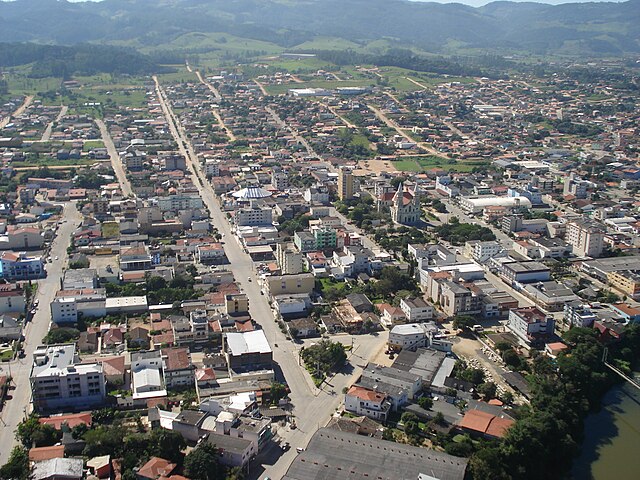 Fotografia aérea em 21 de abril de 2008. No canto inferior direito a ponte pensil sobre o rio Braço do Norte, unindo os bairros Centro e Lado da União. No centro inferior a Praça Coronel Collaço (formato triângulo-retangular) e no meio da foto a Igreja Nosso Senhor do Bonfim, no centro da Praça Padre Roer. No canto superior esquerdo está localizado o bairro Santa Augusta. Nos morros no canto superior esquerdo está o bairro Pinheiral. Fotografia do bioquímico Roberto Pereira, Braço do Norte.