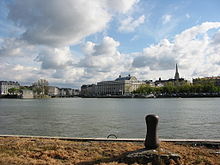 Uitzicht op een rivier, met een kade op de voorgrond, gebouwen op de achtergrond en een blauwe lucht begroeid met stapelwolken.