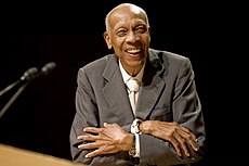 An aged man smiling dressed in a grey suit with his arms crossed.