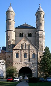 Façade d'église à vaste porche surmonté d'un large et haut fronton triangulaire, devant une massive tour carrée flanquée de deux tours plus élevées, étroites et cylindrique