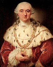A white whigged man with a crimson coat and an ermine mantle holds a medallion in his hand. The medallion has a bright green stone in the center, and is encircled by a star-burst, and it hangs from a jewel-studded chain around his neck.