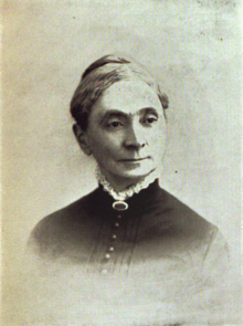 B&W portrait photo of a woman with her hair in an up-do, wearing a dark blouse with white fringe at the collar