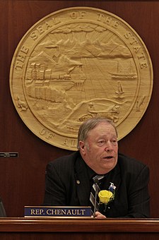 Mike Chenault is the longest-serving speaker, holding the office for four terms from 2009 to 2017. He is shown seated at the speaker's chair at the start of the 29th Legislature in January 2015. A large wood carving depicting the state seal hangs on the wall behind him. Mike Chenault at Speaker's Chair.jpg