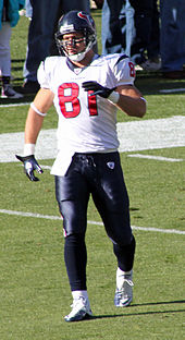 A light-skinned man wearing a white football jersey and dark blue pants