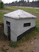 El blanco semienterrado búnker de hormigón plaza con un techo brocha concreto y lagunas horizontales rectangulares. Una valla de altura es visible en el fondo.