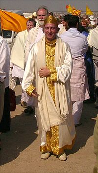 A photograph of a middle-aged caucasian male who dressed in white silk robes with gold trim, wearing a golden crown and standing outdoors in a crowd.