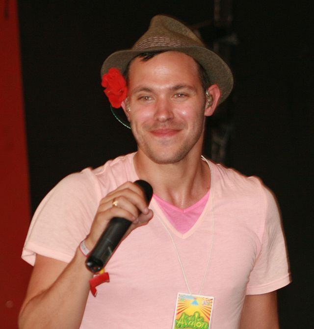 A man with a brown cap wearing a pink T-shirt and holding a microphone. He is looking to the left and smiling.