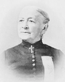 An older white woman, grey hair tightly drawn back, wearing a high-collared dark dress with a row of buttons down the front; she has a cross pin attached at the throat, and a ribbon pinned to her chest