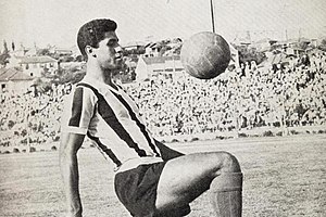 Um homem negro com a camiseta titular do Grêmio fazendo embaixadinha com uma bola num estádio de futebol.