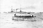 A small boat packed with soldiers passes in front of a cruiser and several transport ships