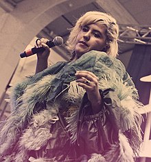 Coco O. is on-stage singing into a microphone and has blonde hair. She wearing a long jacket with feathered fringe.