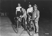 Mustavalkoinen valokuva, jossa kaksi pyöräilijää polkupyörällä, paikallaan radalla, nojaten kahteen siviilivaatteisiin mieheen.