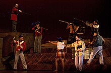 Opera performers are British soldiers on stage pointing rifles down at the main singer and more chorus members who are Māori warriors also with rifles. It is a tense scene.