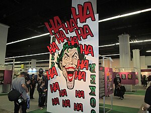 A large playing card bearing the Joker's face stands before a series of art works featuring the Joker