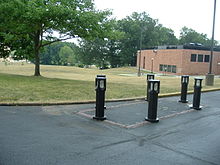 Color photograph of a memorial (six posts with lights set around a rectangular demarcation) with grass, trees, and a building in the background.