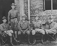 Four men in military uniforms wearing hats. Three are seated on a bench and one is standing behind the others.