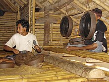 The left front post of a Sumbanese house is visible here complete with the special aperture where women passed rice to be offered to the marapu. Sumba-Gamelan.jpg