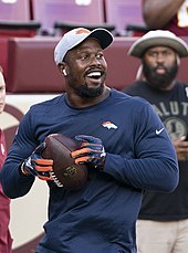 Von Miller con gorra y AirPods sonríe mientras sostiene una pelota de fútbol.  También lleva guantes y parece que pronto lanzará la pelota.