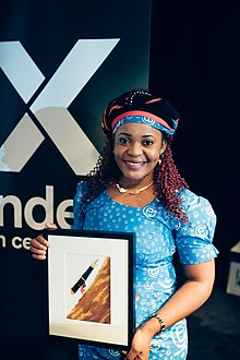 Photograph of Mimi Mefo holding the 2019 Freedom of Expression Journalism Award, a framed artistic painting of her carrying a huge calligraphy pen up a hill