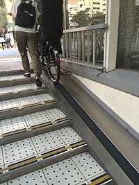 Degraus rasos com uma rampa motorizada de borracha preta deslizante ao lado;  um ciclista empurrando uma bicicleta até o topo da rampa