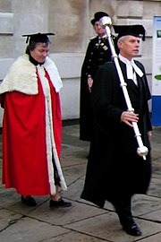 Universitaire portant la robe rouge et le mortar board ou square, sans doute l'invitée d'honneur, précédée du bedell en toge noire et square', avec dans les mains le mace