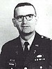 Head and shoulders of a middle-aged man with closely cropped hair and horn-rimmed glasses. He is wearing a dark military jacket with a parachute badge on the left breast and a Christian cross under a "U.S." pin on each lapel.