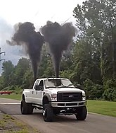 Das Bild zeigt einen Pick-up-Truck, der hinten zwei schwarze Rauchwolken ausstößt.
