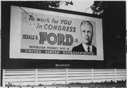 A billboard shows a portrait of a man in a suit, with the text "To work for You in congress" at the top, followed by "Gerald R. Ford Jr.", followed by "Republican Primary September 14", with "United States Representative" across the bottom of the sign.