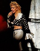Madonna, dressed in a polka-dot blouse and white pants, sings to a microphone she holds to her mouth. Behind here, there's a white structure.