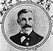 Tête et épaules d'un homme blanc avec une large moustache touffue, portant un nœud papillon et un costume sombre avec une médaille épinglée sur la poitrine gauche.  Le portrait est entouré d'un cadre circulaire et d'images de fleurs.