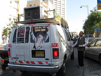 A Na Nach Breslover Hasid stands beside a van emblazoned with images of Rabbi Yisroel Ber Odesser and the "Na Nach Nachma Nachman Meuman" name in downtown Jerusalem. Na Nach truck.jpg