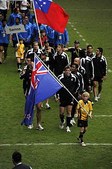 Joueurs de face, défilant en déléguation sur un terrain. Les joueurs sont précédés d'un enfant portant le drapeau de l'équipe.