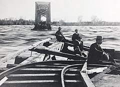 Photo of a flooding river, with a railroad bridge broken in a number of places. Men are sitting and standing on portions of the bridge that aren' damaged.