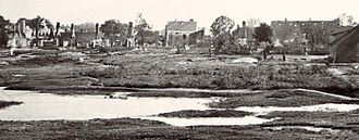 A black and white picture showing a large artillery crater filled with water. In the background a number of ruined burnt-out houses.