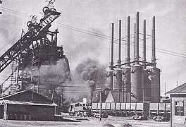 Photo d'une usine prise depuis le sol. D'un bâtiment proche, s'élancent six grandes cheminées.