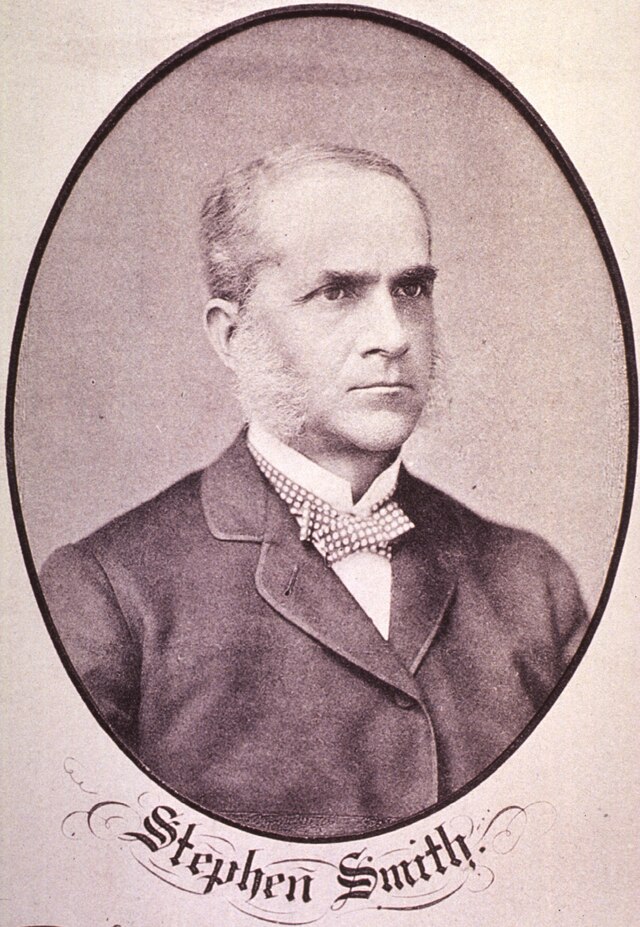 Three-quarters head and shoulders photograph of a middle-aged white man with dark eyebrows and white mutton-cop sideburns wearing a dark coat.