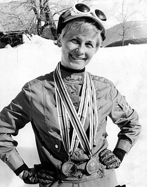 A smiling short-haired blond woman wears a track suit with gloves, and a hat with white skiing goggles. She holds her hands on her waist and carries two medals around her neck. Behind her, a sunny and snowy landscape.