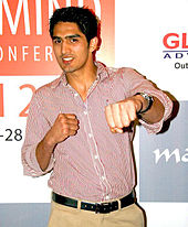 Picture of an young Indian male up to the waist. He has sharp features, short cropped black hair and is clad in a pink striped shirt and khaki pants with a black belt. The man appears to look a little to the right of the camera, smiling and making a gesture with his left hand as if he is punching.