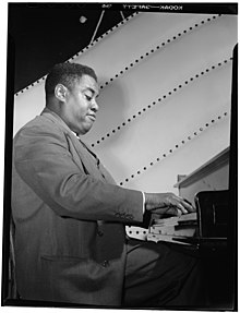 Vu de profil, le pianiste Art Tatum est en train de jouer du piano. Sa main droite effleure les touches, son cors est légèrement en arrière, la tête légèrement inclinée vers le clavier. La photo est en noir et blanc.