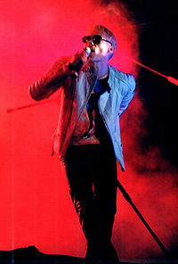 A color photograph of a man on stage at a concert singing in to a microphone with one arm behind his back.